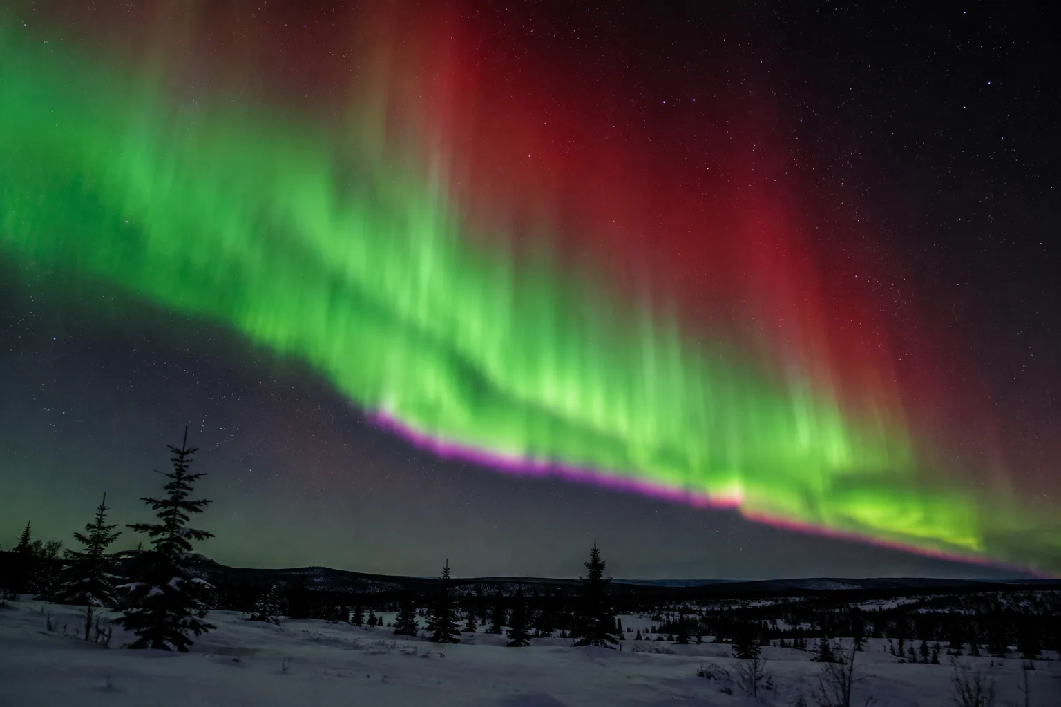 Green auroral curtains with purple fringes and a high red glow sweep above a snowy northern landscape.