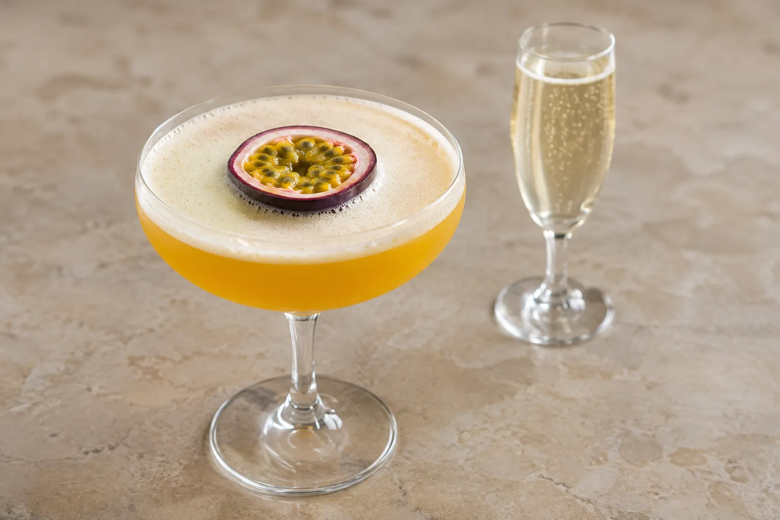 A golden Pornstar Martini topped with a passion fruit half stands beside a small glass of sparkling wine.